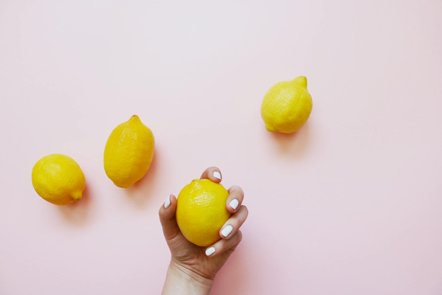 Hand holding a lemon on a soft pink background, surrounded by additional lemons.